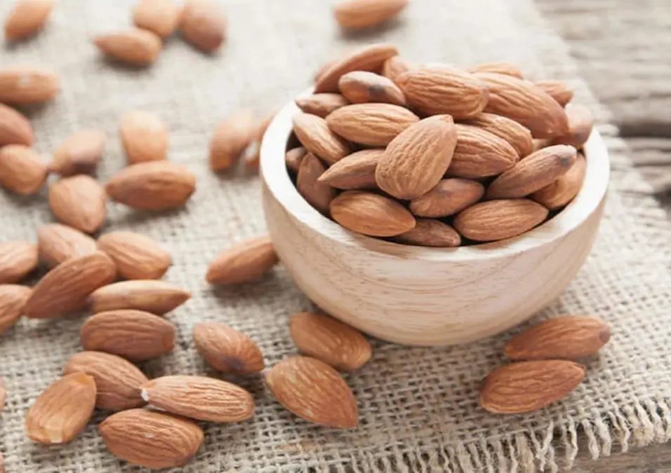وعاء خشبي صغير يفيض باللوز الكامل على سطح من الخيش، مع وجود المزيد من حبات اللوز المتناثرة حوله، مما يوحي بفوائده الصحية العديدة ولماذا يُنصح بتناوله يومياً.