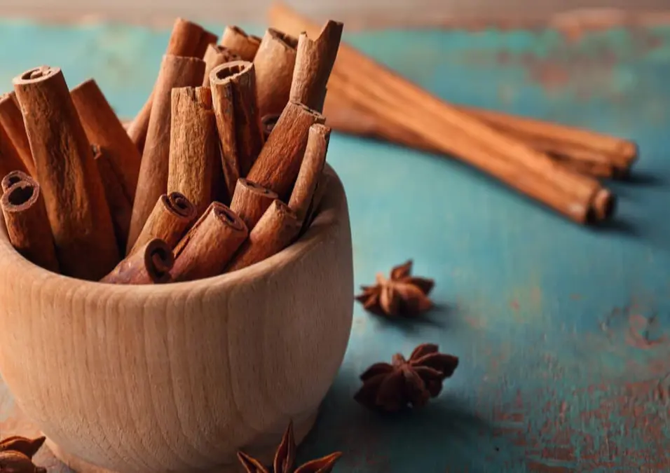 وعاء خشبي مليء بأعواد القرفة على سطح ريفي أزرق، مع وجود يانسون نجمي متناثر حوله، مما يوحي بفوائد القرفة للمعدة والقولون، متى تكون مفيدة ومتى قد تكون مضرة.