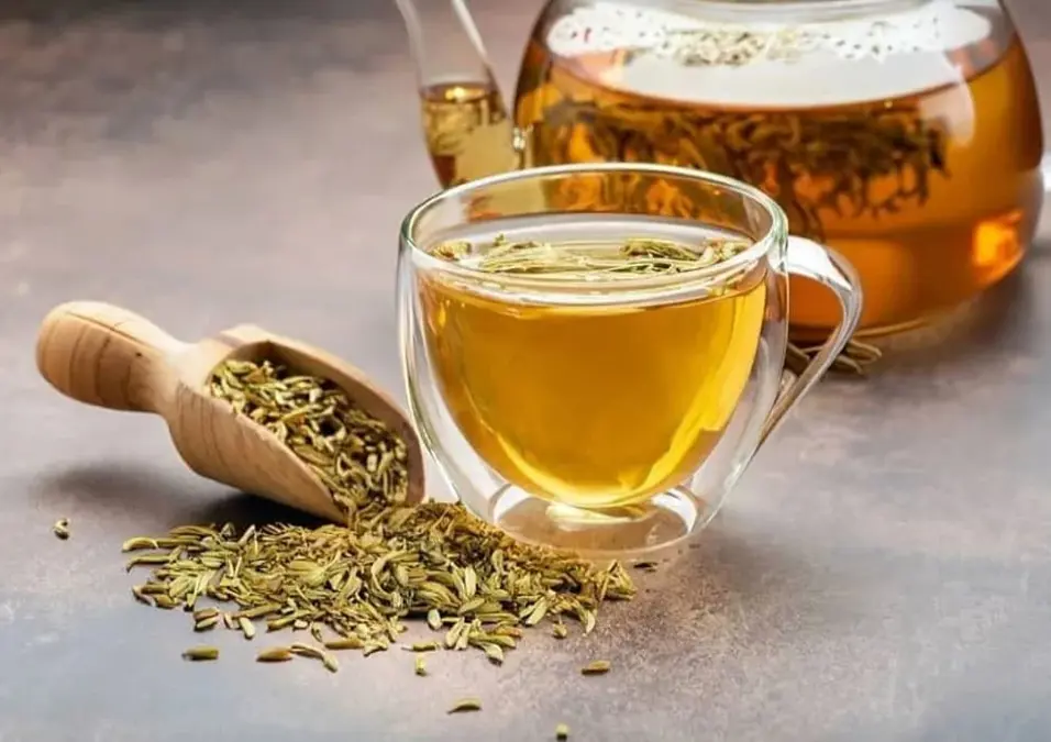 كوب زجاجي وإبريق شاي مملوءان بشاي الشمر، مع مغرفة خشبية من بذور الشمر بالجوار، يوحي بفوائده الصحية وطرق تحضيره.