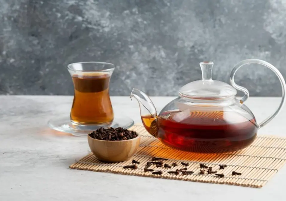 إبريق شاي زجاجي وكوب زجاجي صغير مملوءان بشاي داكن، مع وعاء خشبي من القرنفل بالجوار، مما يوحي بدورها في التنحيف والتخسيس.