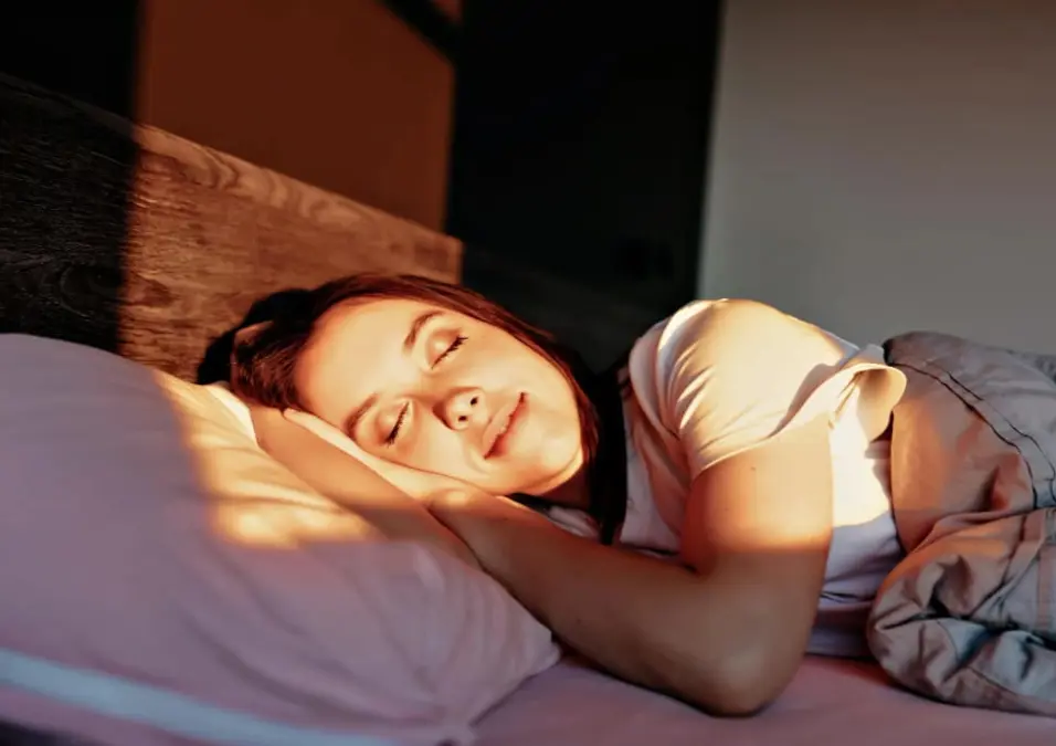 امرأة نائمة بسلام في سريرها وأشعة الشمس تلامس وجهها، مما يوحي بفوائد العصفر للنوم، والتهدئة الطبيعية، وجودة نوم أفضل.