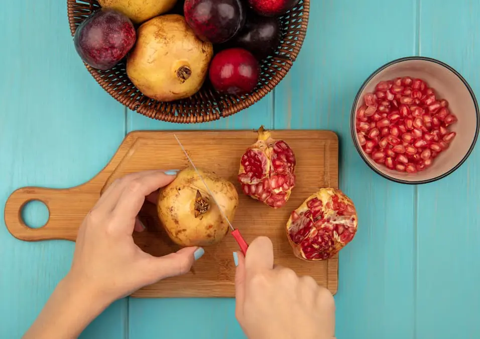 منظر علوي ليدين تقطعان رمانة على لوح خشبي، مع الحبوب وفواكه أخرى بالجوار، تستكشف فوائد الرمان للسكري.