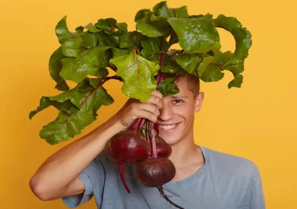 شاب مبتسم يحمل حزمة من الشمندر الطازج مع أوراق خضراء كبيرة أمام وجهه، مما يوحي بفوائد الخضار للطاقة والأداء البدني للرجال.
