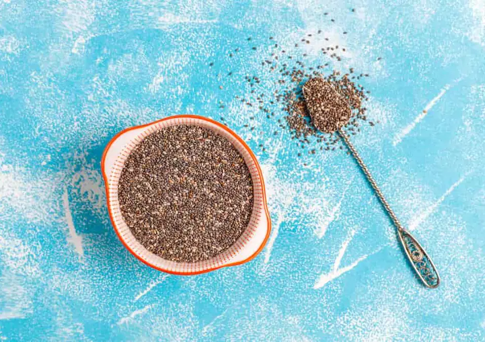 وعاء وملعقة مليئة ببذور الشيا على خلفية زرقاء، تمثل فوائد بذور الشيا للنساء ككنز طبيعي للصحة، الجمال، والخصوبة.