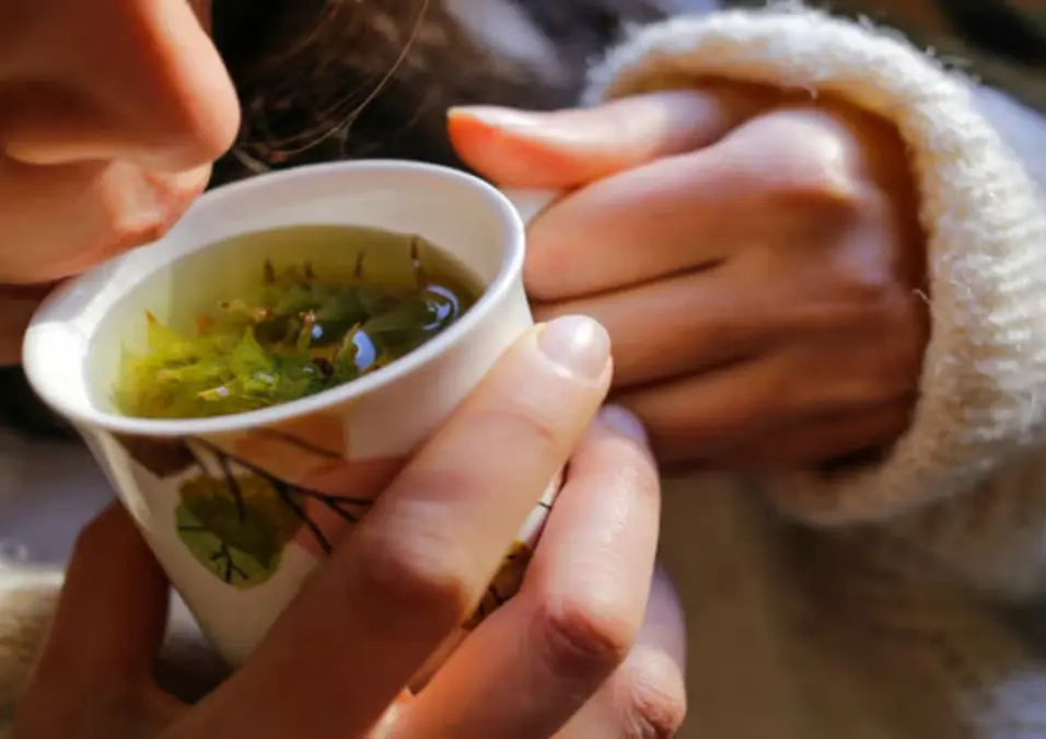 لقطة قريبة ليدي امرأة تمسكان بكوب سيراميك مزخرف وهي ترتشف شاي الميرمية الدافئ مع ظهور الأوراق بداخله، مما يوحي بفوائد الميرمية للنساء.