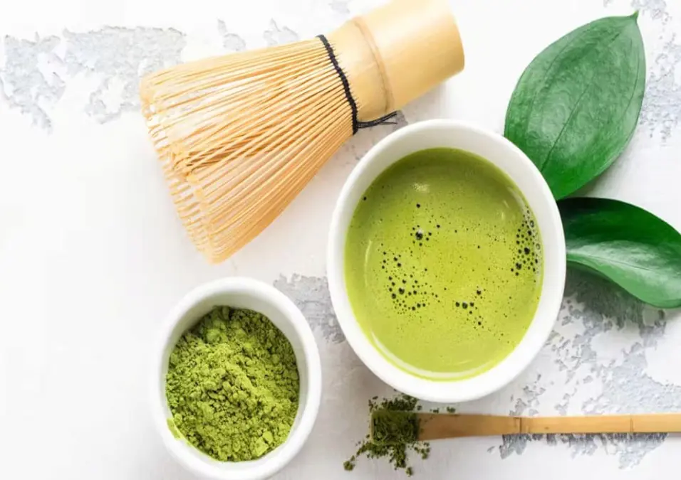 وعاء من شاي الماتشا الجاهز مع مضرب ومسحوق الماتشا على خلفية بيضاء، يوحي بفوائد الماتشا لصحة الجسم والعقل.