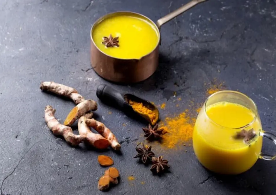 قدر نحاسي صغير وكوب زجاجي مملوءان بـ حليب الكركم الذهبي على سطح حجري داكن، بجانب جذور الكركم الطازجة واليانسون النجمي، مما يتساءل عن فوائد الكركم على الريق وهل الاستمرار عليه آمن.