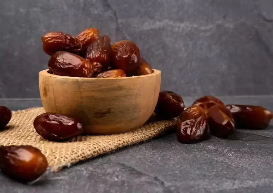 وعاء خشبي يفيض بـ التمر الناضج على قطعة من الخيش، مع حبات تمر إضافية متناثرة حوله على خلفية حجرية داكنة، مما يسلط الضوء على فوائد التمر للجسم وأفضل وقت لتناوله.