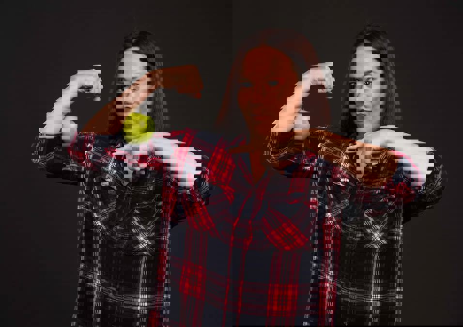 امرأة تستعرض عضلة ذراعها مع تفاحة عليها، توضح كيفية بناء عضلات قوية بشكل طبيعي بدون مكملات غذائية.
