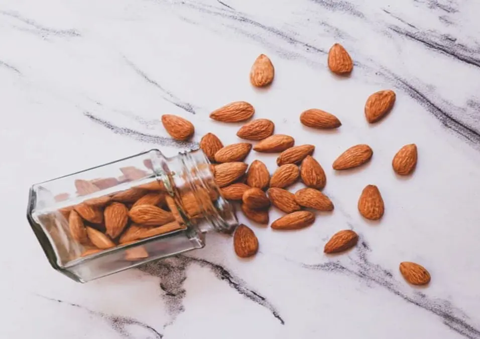 مرطبان زجاجي ملقى على جانبه على سطح رخامي أبيض مع لوز كامل يتناثر منه، مما يتساءل عن فوائد اللوز لمرضى السكري وهل يرفع السكر أم يساعد على استقراره.