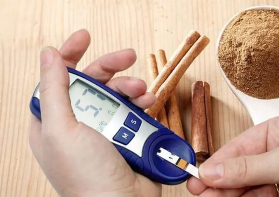 يدان تستخدمان جهاز قياس نسبة السكر في الدم، مع وجود أعواد قرفة ووعاء من القرفة المطحونة في الخلفية، مما يتساءل عن فوائد القرفة لمرضى السكري والحقيقة وراء ذلك.