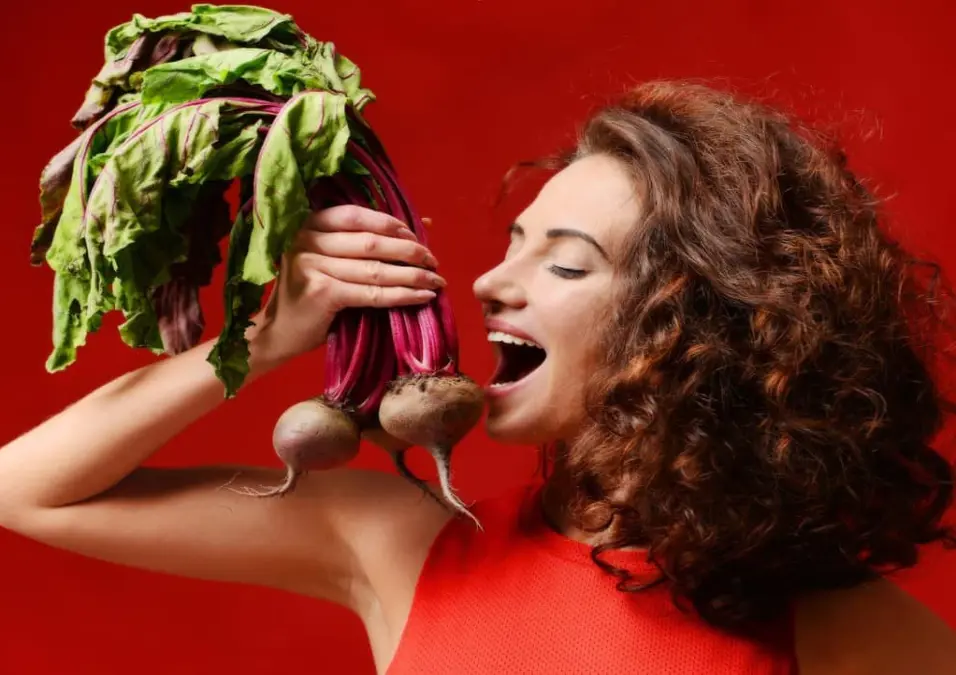 امرأة ذات شعر مجعد تحمل حزمة من الشمندر الطازج بالقرب من وجهها على خلفية حمراء، توحي بفوائد الشمندر للنساء في دعم الهرمونات والدورة الشهرية.