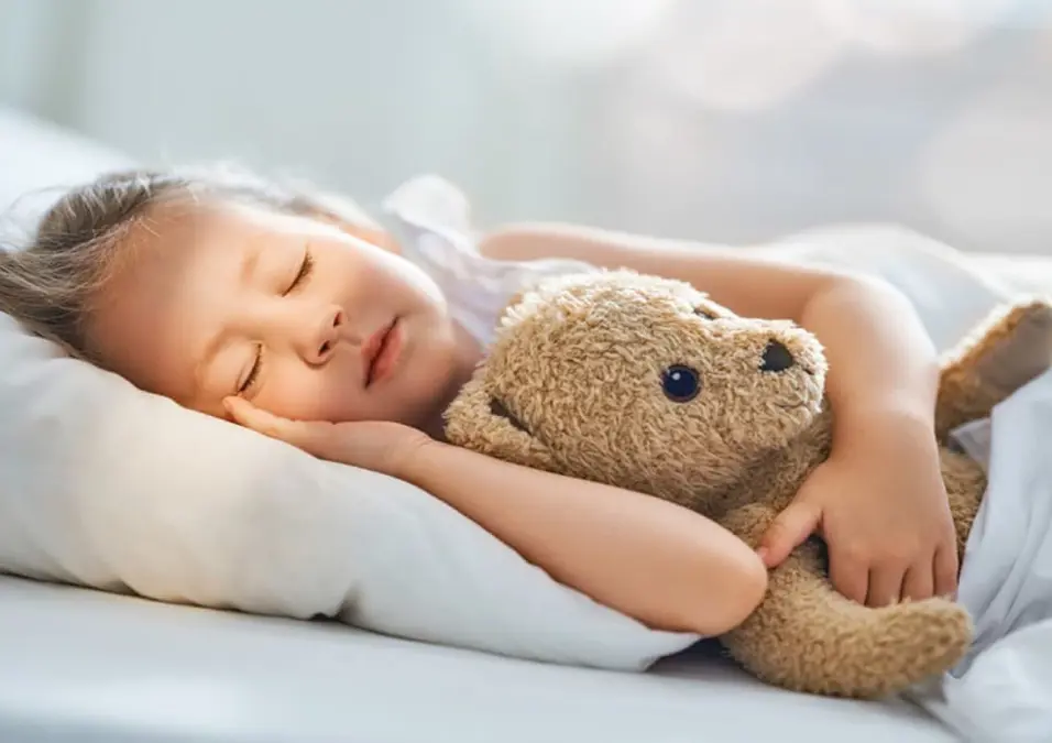 طفل صغير ينام بعمق في سرير أبيض وهو يعانق دمية دب، مما يوحي بفوائد العصفر للأطفال في المساعدة على النوم والتهدئة.