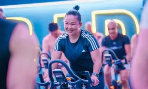 GNSmiling woman in a LES MILLS SPRINT class at GymNation, riding an indoor bike with neon lights in the background.