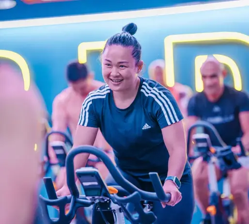 Smiling woman in a LES MILLS SPRINT class at GymNation, riding an indoor bike with neon lights in the background.
