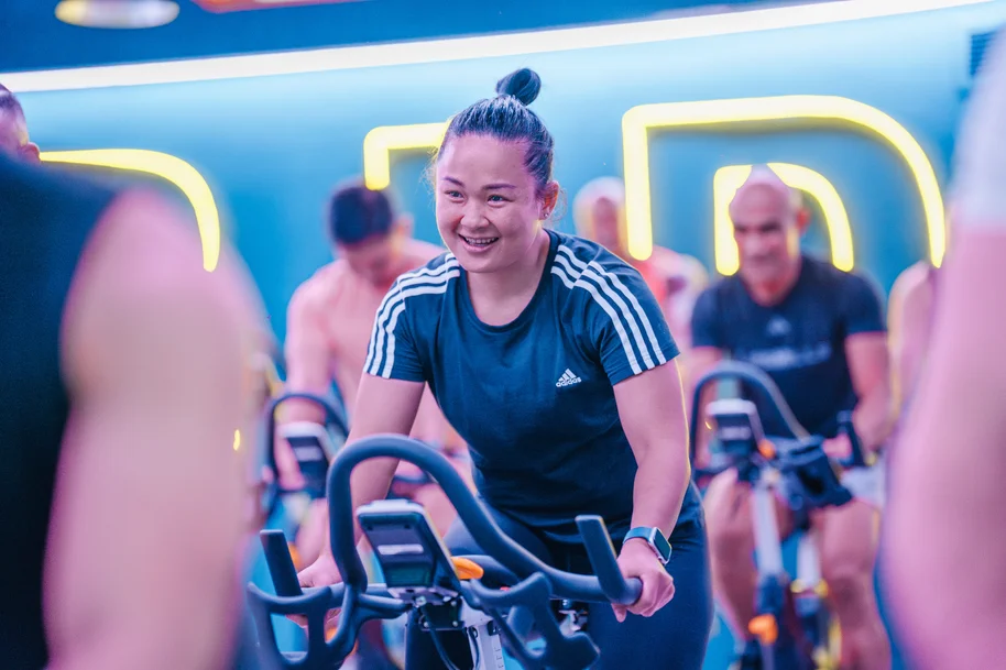 Smiling woman in a LES MILLS SPRINT class at GymNation, riding an indoor bike with neon lights in the background.