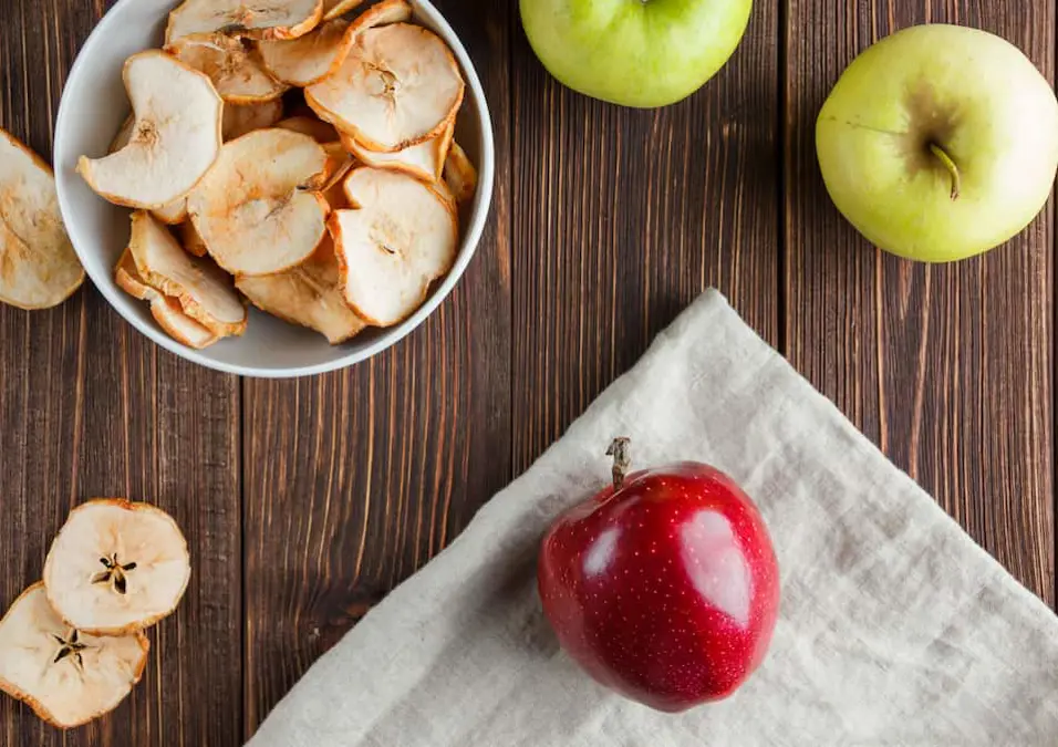  تفاحة حمراء على منديل مع شرائح تفاح مجففة وتفاح أخضر، تستكشف نسبة الكربوهيدرات في التفاح الأحمر.