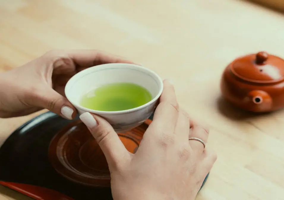 يدان تحملان كوبًا صغيرًا من الشاي الأخضر مع إبريق شاي على طاولة خشبية، تمثل تجربة شخصية مع الشاي الأخضر للقولون والتنحيف.