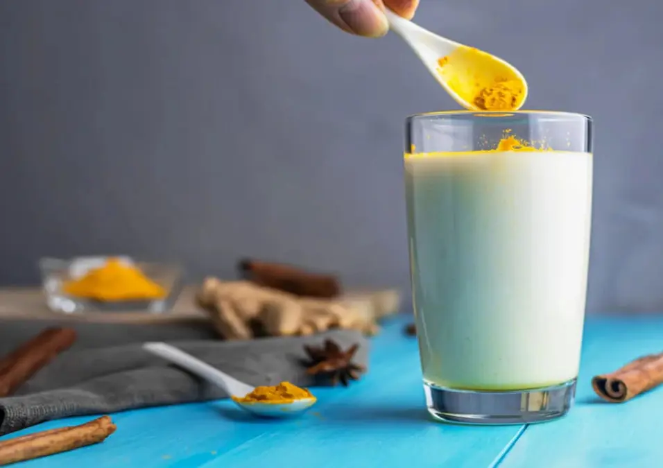 يد تضيف ملعقة من مسحوق الكركم الزاهي إلى كوب طويل من الحليب الأبيض، مما يوحي بفوائد الكركم مع الحليب كـ 