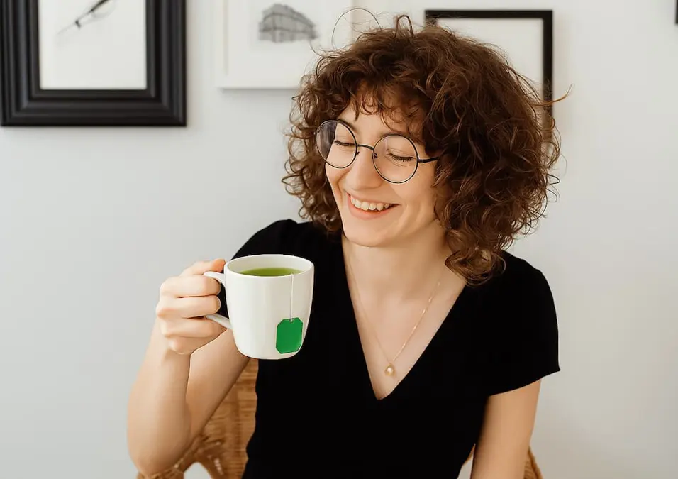 امرأة تبتسم وهي تحمل كوبًا من الشاي الأخضر، يوحي بفوائد الشاي الأخضر كسر لجمال وصحة المرأة.