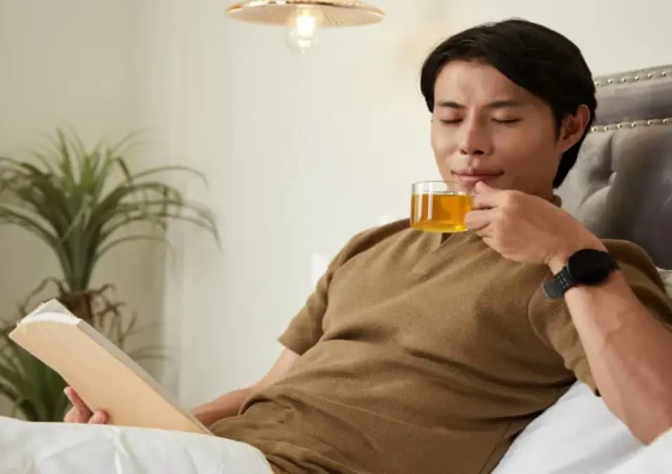 رجل يسترخي في السرير، يحتسي الشاي ويقرأ كتاباً، يوحي بفوائد البابونج للرجال المثبتة علمياً.