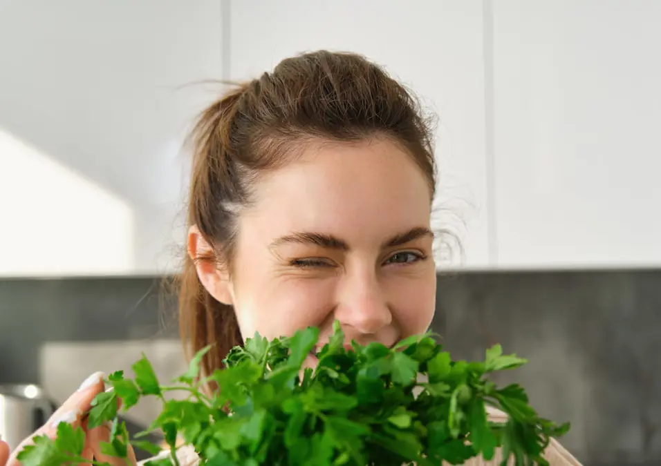 امرأة تغمز وتبتسم وهي تحمل حزمة من الجرجير (أو خضروات مشابهة) على وجهها، توحي بفوائد الجرجير للحامل في مراحل الحمل المختلفة.