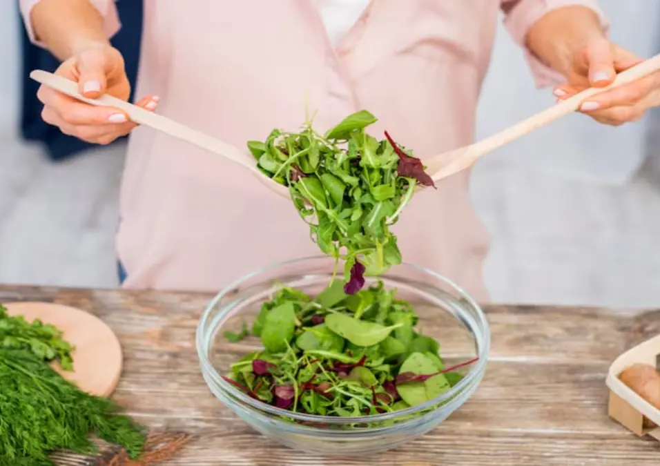 امرأة تقلب سلطة تحتوي على الجرجير وخضروات أخرى في وعاء زجاجي، توحي بفوائد الجرجير للنساء.