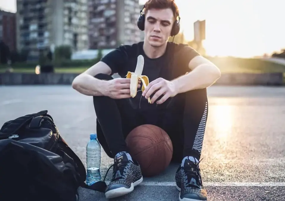 رجل يرتدي ملابس رياضية يأكل موزة في الهواء الطلق بجانب كرة سلة، يوحي بفوائد الموز للعضلات ولماذا يوصي به الخبراء.