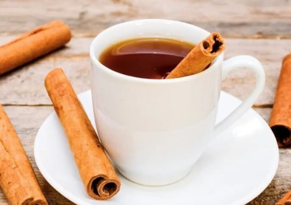 كوب أبيض من شاي القرفة بداخله عود قرفة، موضوع على سطح خشبي ومحاط بالمزيد من الأعواد، مما يوحي بفوائد القرفة قبل النوم في التنحيف وتحسين جودة النوم.