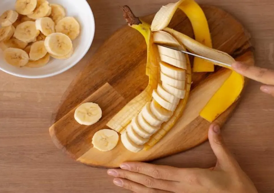 يدان تقطعان موزة على لوح خشبي، مع شرائح مقطعة بالجوار، توحي بفوائد الموز على الريق.