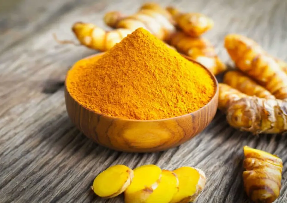 وعاء خشبي صغير مملوء بـ مسحوق الكركم الزاهي، محاط بجذور وشرائح الكركم الطازجة على سطح خشبي ريفي، مما يتساءل عن دوره في التنحيف وإنقاص الوزن.