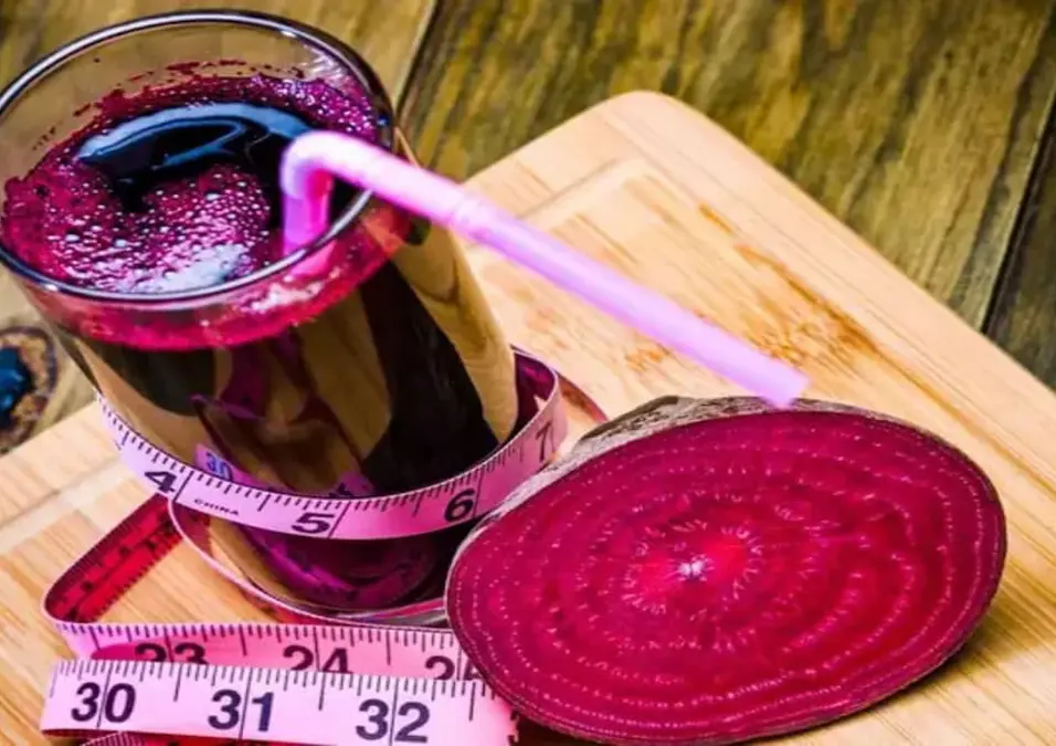 كوب من عصير الشمندر الداكن مع ماصة وشمندر مقطع على لوح خشبي، يحيط به شريط قياس وردي، مما يوحي بدور الشمندر في التنحيف والتخسيس.