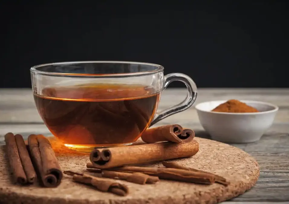 كوب زجاجي من شاي القرفة على قاعدة فلينية، محاط بأعواد القرفة ووعاء صغير من القرفة المطحونة، مما يوحي بفوائد شرب القرفة يومياً، وأفضل وقت لتناولها والكمية الآمنة.