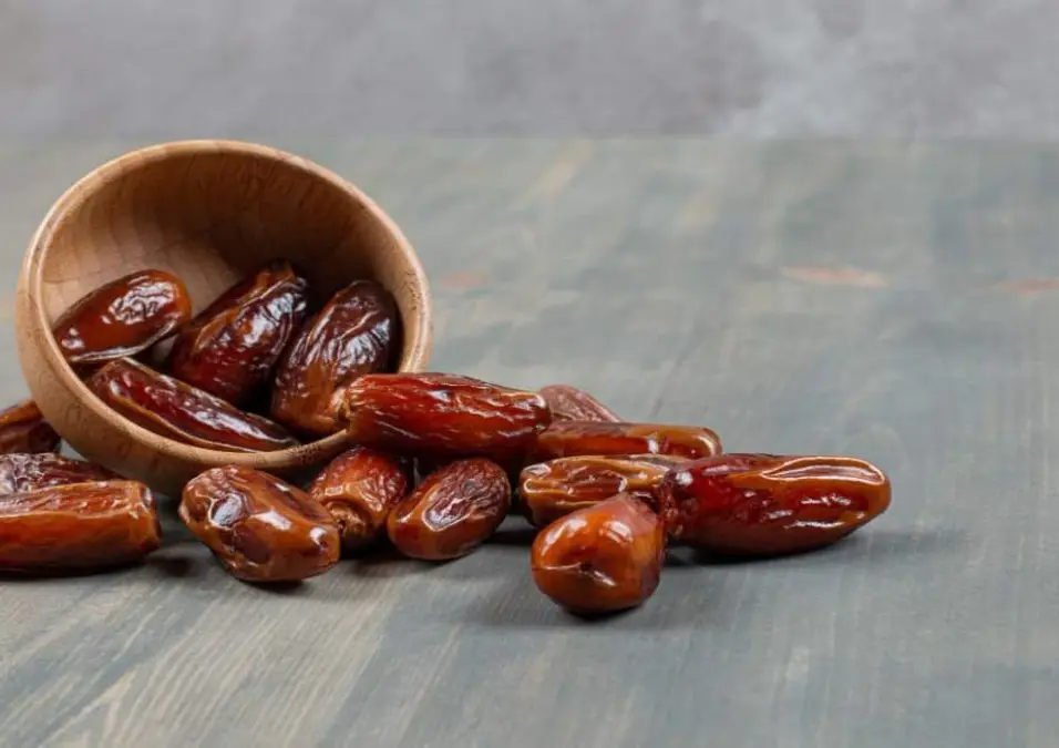 وعاء خشبي صغير مائل على جانبه على سطح خشبي ريفي مع تمور بنية ناضجة تتساقط منه، مما يتساءل عن فوائد التمر للرجيم وكم حبة يمكن تناولها يومياً.
