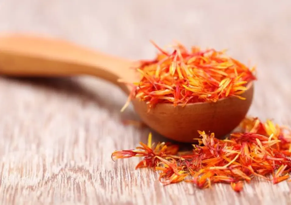 ملعقة خشبية صغيرة تفيض بتويجات العصفر المجففة ذات اللون البرتقالي الزاهي على سطح خشبي، مما يوحي بفوائده للأعصاب.