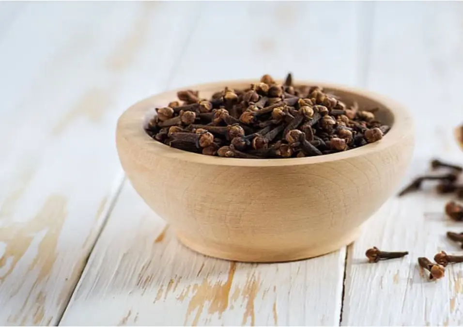 وعاء خشبي صغير مليء بالقرنفل المجفف على سطح خشبي أبيض، مما يوحي بفوائد القرنفل للشعر وكيفية استخدامه.