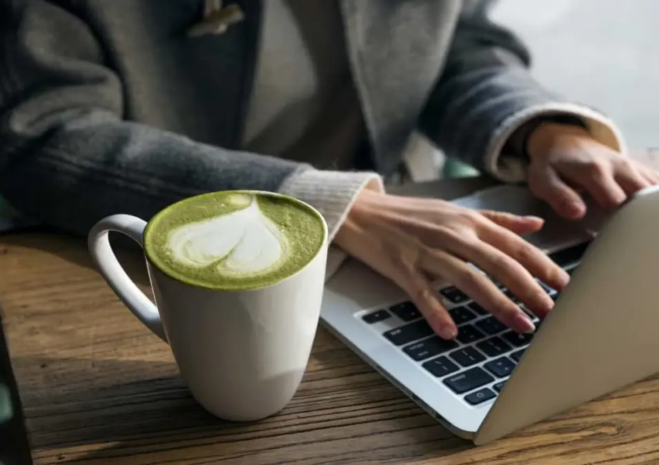 كوب من ماتشا لاتيه بتصميم قلب على طاولة خشبية، بجانب شخص يستخدم حاسوبًا محمولًا، يوحي بالماتشا كمشروب نحو صحة أفضل للرجال.