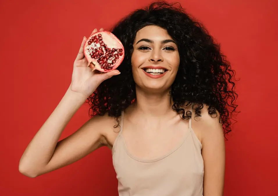 امرأة ذات شعر مجعد تبتسم وتحمل نصف رمانة، توحي بسر الجمال والصحة للمرأة.