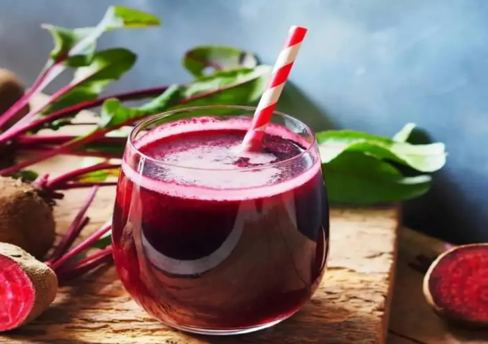 كوب من عصير الشمندر الزاهي مع ماصة باللونين الأحمر والأبيض، يظهر في الخلفية حبات شمندر طازجة مع أوراقها الخضراء، مما يوحي بفوائده الصحية وما يحدث للجسم عند شربه.