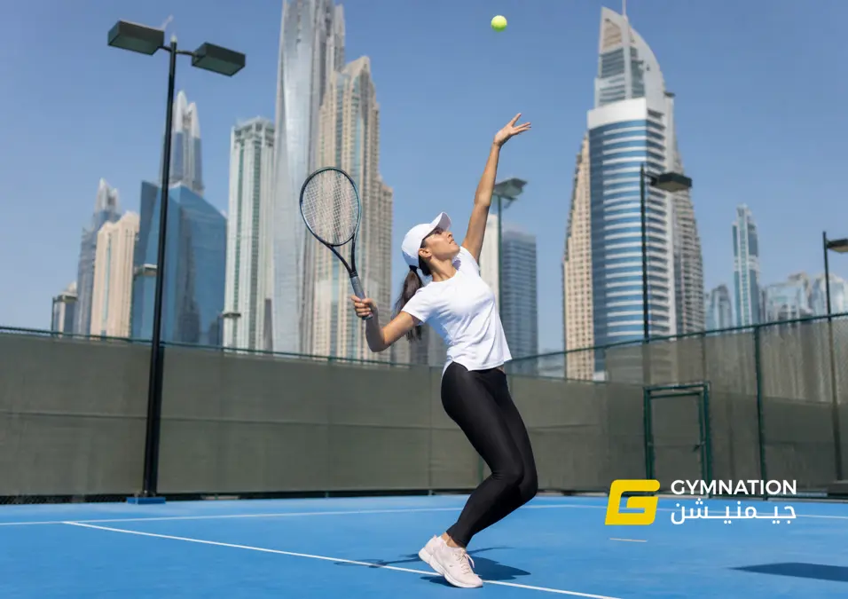 A player serves on a bright blue court, with skyscrapers in the background. Sunlight casts sharp shadows. The atmosphere