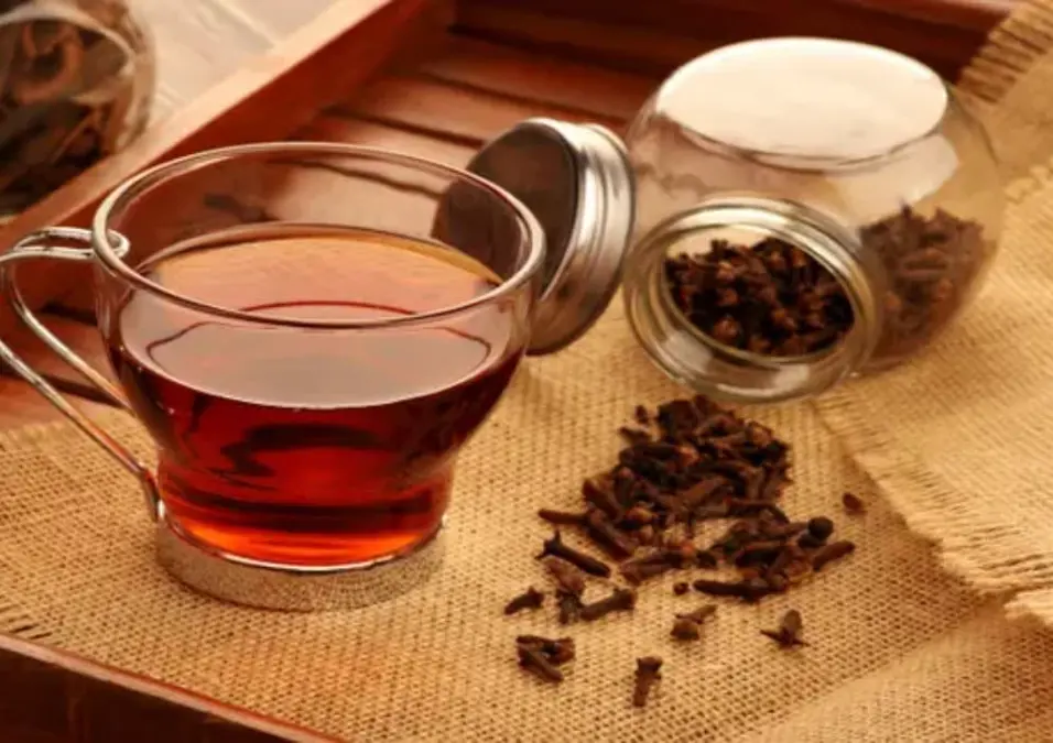 كوب زجاجي من مغلي القرنفل الداكن على سطح من الخيش، بجانب مرطبان زجاجي مليء بالقرنفل، مما يوحي بفوائد القرنفل المغلي وطريقة استخدامه الصحيحة ومحاذيره.