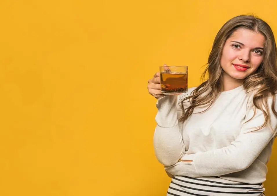 امرأة مبتسمة تحمل كوباً زجاجياً من الشاي الداكن أمام خلفية صفراء زاهية، مما يوحي بفوائد القرنفل للنساء، وتحديداً فيما يتعلق بالهرمونات والرحم والبشرة.